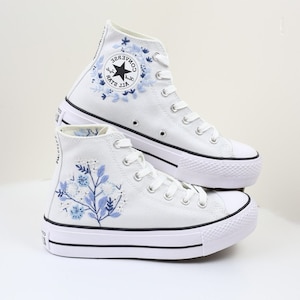 May include: White high-top Converse sneakers with a platform sole and blue floral embroidery. The Converse logo is visible on the side of the shoe.