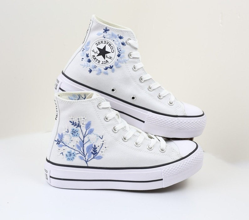 May include: White high-top Converse sneakers with a platform sole and blue floral embroidery. The Converse logo is visible on the side of the shoe.