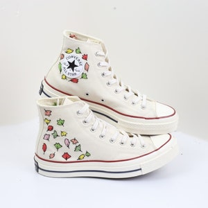 K&ouml;nnte beinhalten: Wei&szlig;e Converse-Sneaker mit hohem Schaft und einem Muster aus roten, gelben und gr&uuml;nen Bl&auml;ttern. Die Schuhe haben ein schwarzes Sternlogo und den Schriftzug "Converse All Star" an der Seite.