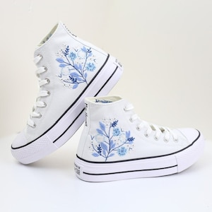 May include: White high-top canvas sneakers with blue floral embroidery. The sneakers have a platform sole and white laces.