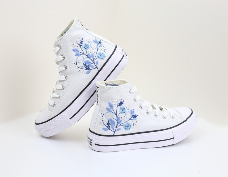 May include: White high-top canvas sneakers with blue floral embroidery. The sneakers have a platform sole and white laces.
