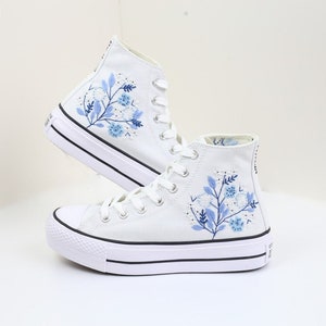 May include: White high-top canvas sneakers with blue floral embroidery. The shoes have a thick white sole and black trim.