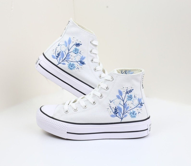 May include: White high-top canvas sneakers with blue floral embroidery. The shoes have a thick white sole and black trim.