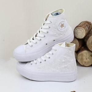 May include: Pair of white high-top sneakers with floral embroidery. The sneakers feature white laces, a rubber toe cap, and a star logo on the side. The shoes are made of canvas material.