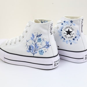 May include: White high-top Converse sneakers with a platform sole. The shoes feature blue floral embroidery and the text "Mrs. Howell" on the side. The Converse All Star logo is visible on the side of the shoe.