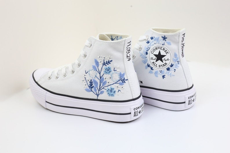 May include: White high-top Converse sneakers with a platform sole. The shoes feature blue floral embroidery and the text "Mrs. Howell" on the side. The Converse All Star logo is visible on the side of the shoe.