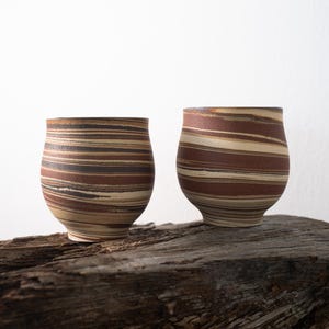 Op de afbeelding: Twee handgemaakte keramische bekers met een uniek streepjespatroon in bruin, beige en zwart. De bekers hebben een afgeronde vorm en staan op een verweerd houten oppervlak. De bekers zijn ongeveer 10 cm hoog.