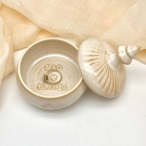 May include: A white ceramic ring box with a lid. The box has a raised design and a knob on the top. The inside of the box is inscribed with "M&G" and "19.10.23". A gold ring is inside the box.