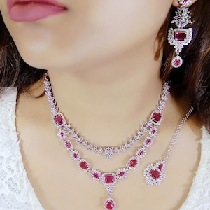 Peut inclure: Un ensemble de bijoux éblouissant comprenant un collier, des boucles d'oreilles et un bijou de tête. L'ensemble est argenté et orné de pierres précieuses rouges rectangulaires et rondes vibrantes, complétées par de plus petites pierres claires. Le collier est à plusieurs rangs.