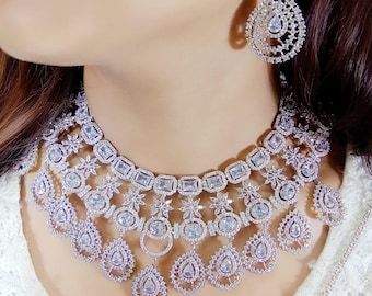 May include: A silver-coloured jewellery set comprising a necklace, earrings, and a headpiece. The necklace features rectangular and teardrop-shaped pendants, embellished with clear gemstones. The earrings are teardrop-shaped, and the headpiece has a similar design.