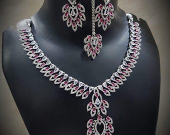 Peut inclure: Un ensemble de bijoux argentés comprenant un collier, des boucles d'oreilles et un bijou de tête. L'ensemble est orné de pierres précieuses claires et roses disposées en forme de feuilles. Le collier et les boucles d'oreilles sont exposés sur une tête de mannequin noire.