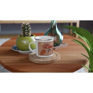 May include: White enamel mug with a silver rim featuring a fall-themed design. The design includes a mushroom, pumpkin, and the word "Welcome" in a fall color palette. The mug sits on a wooden coaster.