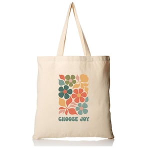May include: A cream-colored canvas tote bag featuring two long shoulder straps. The front of the bag displays a graphic print of stylized flowers in shades of orange, green, yellow, and blue, with the text CHOOSE JOY printed in blue below.