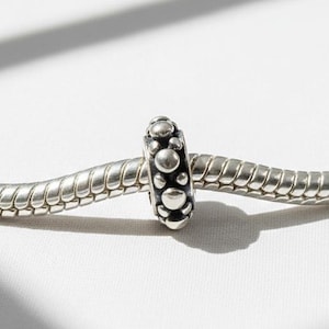 May include: A silver charm bead with a pattern of small, raised circles. The bead is on a silver snake chain, set against a white background with shadows.