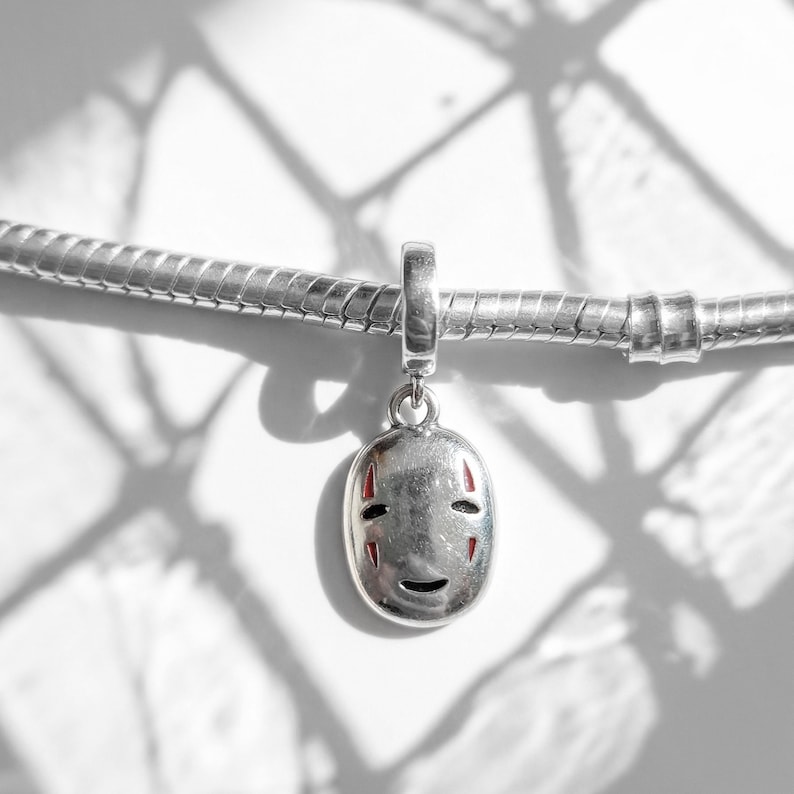 Pu&ograve; includere: Un charm d'argento raffigurante un volto stilizzato con occhi neri e bocca sorridente, con accenti rossi. Il charm &egrave; sospeso a una catena d'argento, su sfondo bianco con ombre.