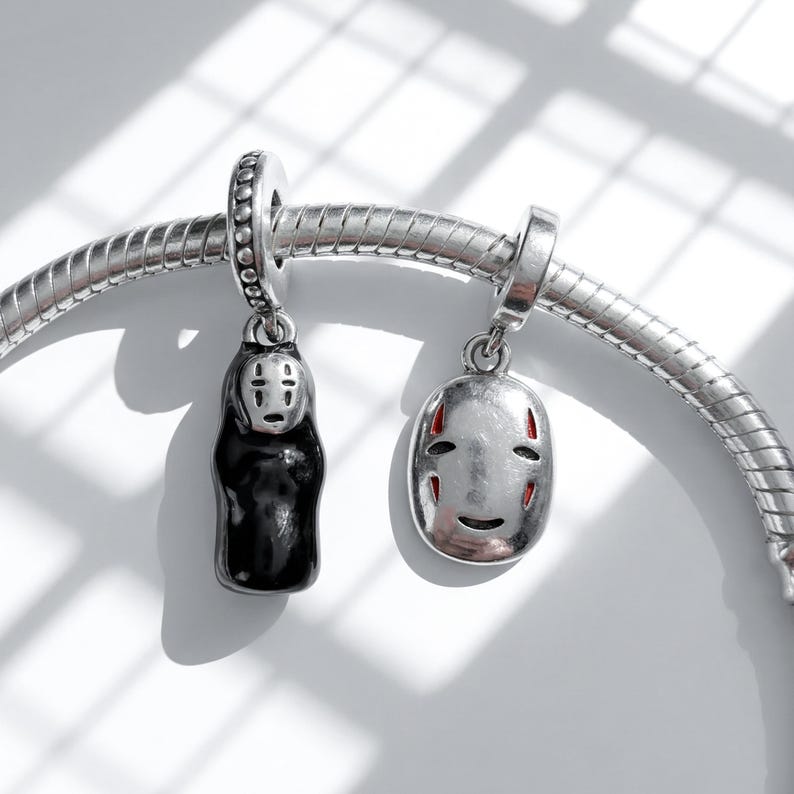 Pu&ograve; includere: Due ciondoli d'argento raffiguranti personaggi del film d'animazione La citt&agrave; incantata. Uno &egrave; una figura nera con un volto bianco, l'altro una maschera d'argento con dettagli rossi. Entrambi sono attaccati a un braccialetto d'argento.