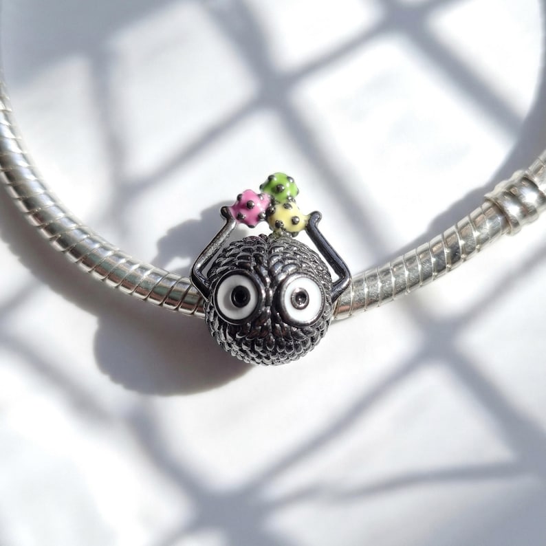 Pu&ograve; includere: Un charm d'argento con un corpo sferico nero strutturato e grandi occhi bordati di bianco. Due bracci neri si estendono verso l'alto, tenendo tre accenti di perline colorate in rosa, verde e giallo. Il charm &egrave; su un braccialetto d'argento.