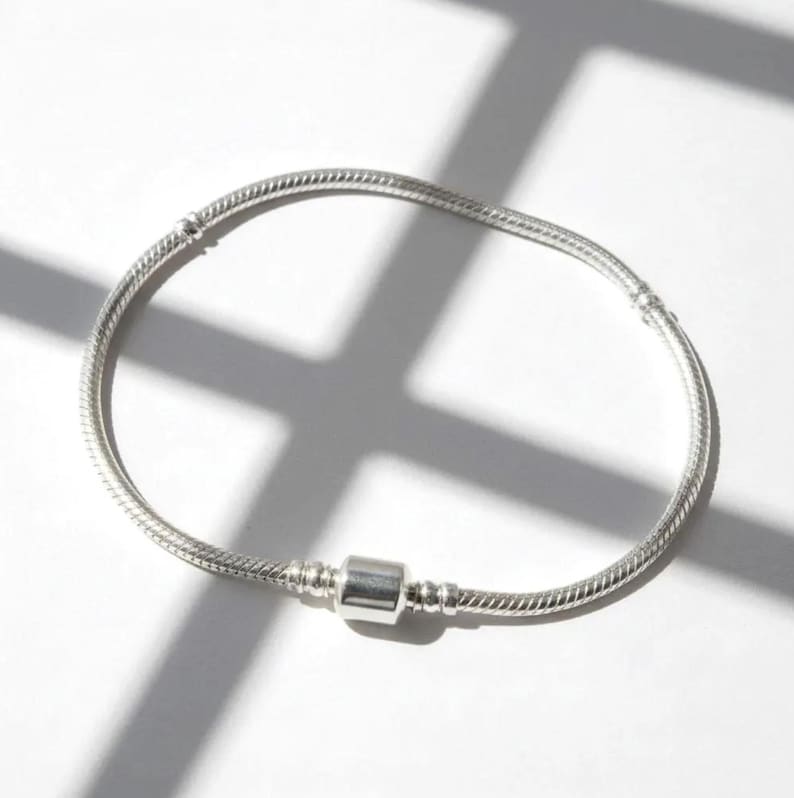 Pu&ograve; includere: Un braccialetto d'argento con un design a catena a serpente e una chiusura a barilotto. Il braccialetto &egrave; realizzato in un materiale metallico lucido ed &egrave; esposto su una superficie bianca con ombre.