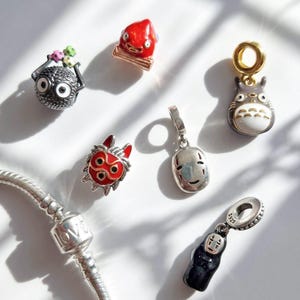 Pu&ograve; includere: Una collezione di ciondoli in argento con personaggi di film d'animazione. I ciondoli includono una creatura rotonda e nera con grandi occhi, una figura rossa, un Totoro grigio e un ciondolo d'argento con la parola "LOVE".