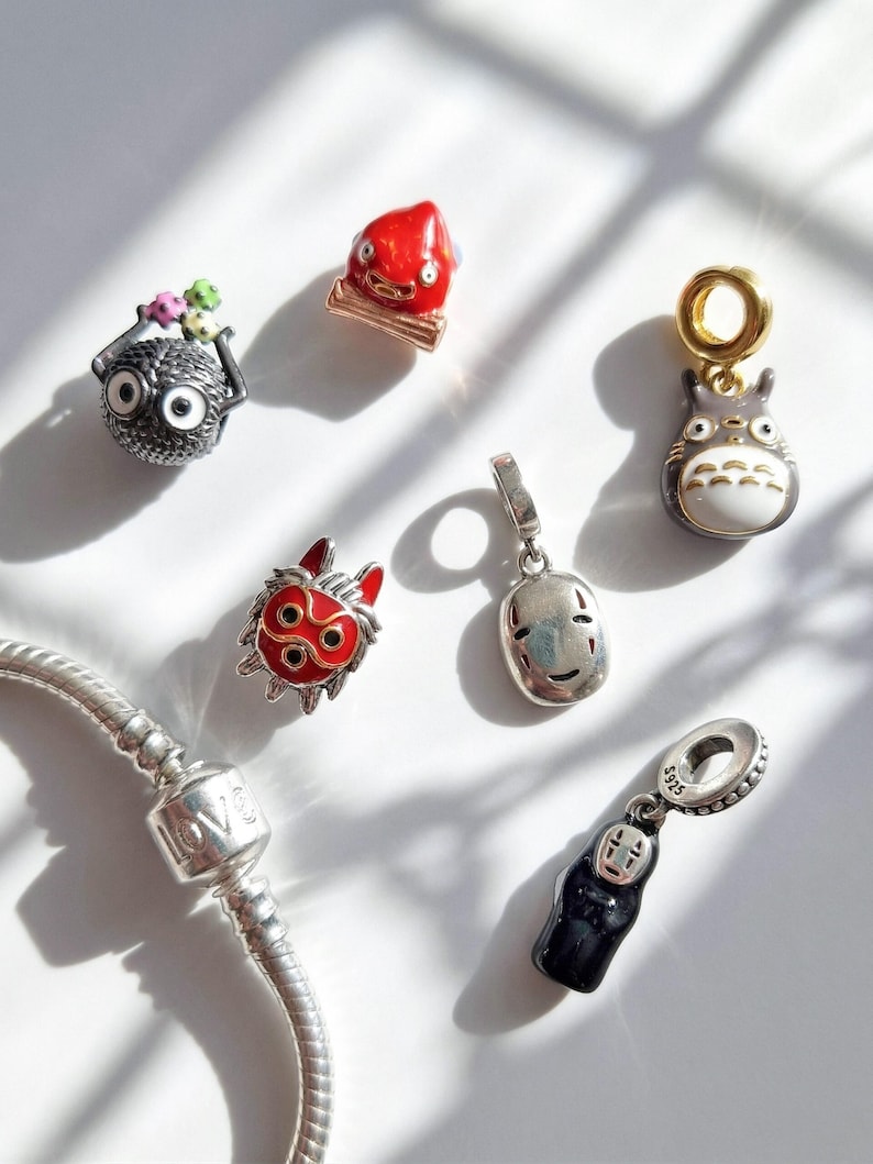 Pu&ograve; includere: Una collezione di ciondoli in argento con personaggi di film d'animazione. I ciondoli includono una creatura rotonda e nera con grandi occhi, una figura rossa, un Totoro grigio e un ciondolo d'argento con la parola "LOVE".