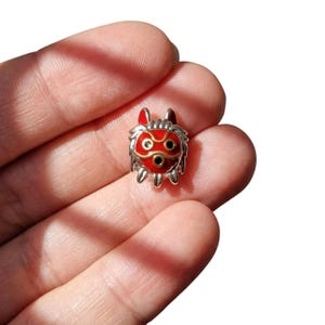 Pu&ograve; includere: Un piccolo ciondolo argentato raffigurante una maschera rossa con dettagli neri e dorati. La maschera presenta un design dettagliato, probabilmente un ciondolo o un charm per gioielli o accessori.