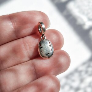 Pu&ograve; includere: Charm color argento con un design Senza Volto. Lo charm ha un piccolo anello nella parte superiore per il fissaggio a un braccialetto o una collana. Il viso ha occhi e bocca ritagliati. Lo charm &egrave; tenuto in mano su sfondo bianco.