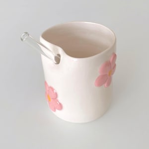 May include: A white ceramic mug with a clear glass stirring rod. The mug features three pink flower embellishments with yellow centers. The mug has a unique spout design for easy pouring.