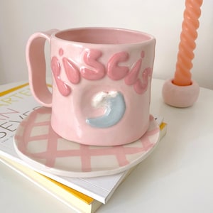 Op de afbeelding: Een roze keramische mok en schotelset. De mok heeft het woord "Pisces" in verhoogde roze letters en een blauw maansikkelontwerp. De schotel heeft een roze en wit geblokt patroon. Een roze kaars is ook zichtbaar.