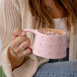 May include: A pink ceramic mug with a handle, filled with a beverage topped with foam and sprinkles. The mug features a grid pattern with small pink hearts. The mug is being held by a person wearing a cream-colored sweater.