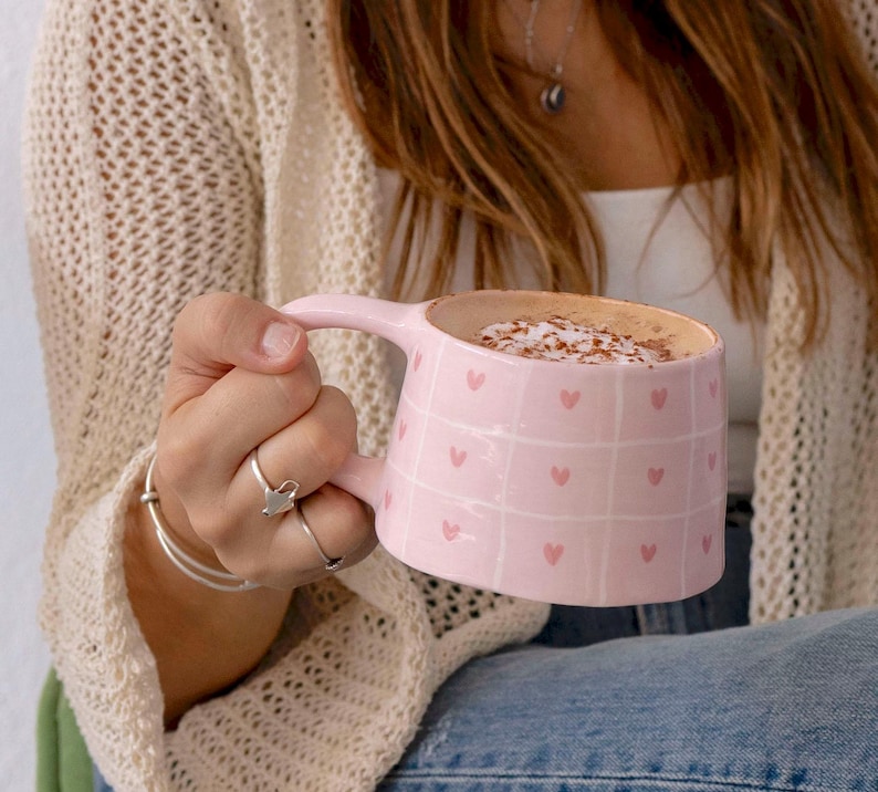 May include: A pink ceramic mug with a handle, filled with a beverage topped with foam and sprinkles. The mug features a grid pattern with small pink hearts. The mug is being held by a person wearing a cream-colored sweater.
