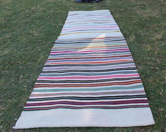 May include: A long, white, woven rug with colorful stripes. The rug is made of natural fibers and has a rustic, handmade look.