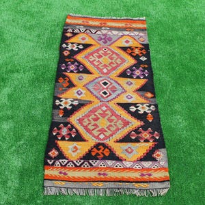 May include: A rectangular, hand-woven rug with a geometric pattern. The rug features a black background with orange, red, purple, and gray diamond shapes. The edges are finished with fringe.