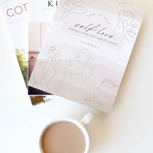 May include: A white journal with the title "The 30 Day Self-Love Journal: Falling in Love with Myself (Again)" and line drawings of faces. The journal is on a white surface with a cup of coffee.