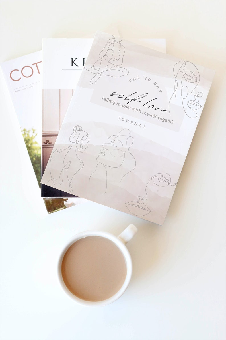 May include: A white journal with the title "The 30 Day Self-Love Journal: Falling in Love with Myself (Again)" and line drawings of faces. The journal is on a white surface with a cup of coffee.