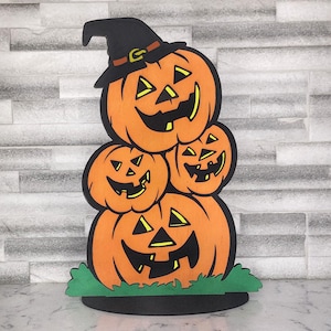May include: A stack of four orange pumpkins with black and yellow painted faces. The top pumpkin is wearing a black witch hat with a brown band. The pumpkins are sitting on a green grass base.