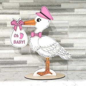 Puede incluir: Una figura de madera de cigüeña rosa y blanca que sostiene una bolsa de regalo para bebé rosa y blanca con el texto "Oh Baby!" La cigüeña lleva un sombrero rosa y un lazo rosa.
