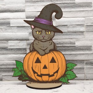 Könnte beinhalten: Eine hölzerne Halloween-Dekoration mit einer grauen Katze, die einen schwarzen Hexenhut mit einem violetten Band trägt und auf einem orangefarbenen Kürbis mit einem geschnitzten Jack-o'-Lantern-Gesicht sitzt.