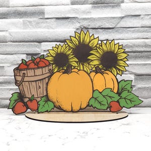 Könnte beinhalten: Eine hölzerne Herbst-Tischdekoration mit drei Kürbissen, einem Korb mit roten Äpfeln und drei Sonnenblumen mit gelben Blütenblättern und braunen Zentren.