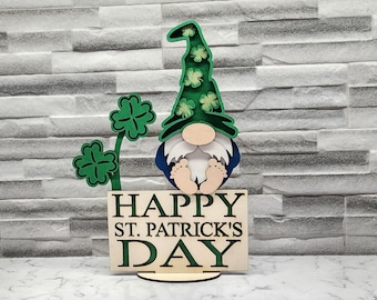 May include: A wooden gnome figurine with a green hat decorated with shamrocks. The gnome is holding a sign that reads "Happy St. Patrick's Day".