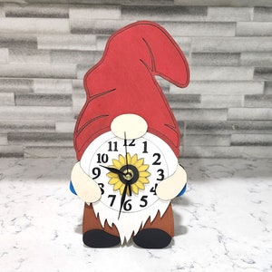 May include: A wooden gnome figurine with a red hat and a white clock face featuring a sunflower design. The clock face has black numbers and hands.