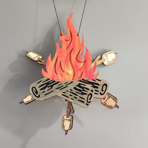 May include: A wooden cutout of a campfire with a red and orange flame, two logs, and four marshmallows on sticks. The campfire is hanging from the ceiling.