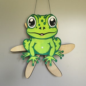 Puede incluir: Una rana verde con ojos grandes y una sonrisa burlona está unida a un móvil de madera en forma de estrella. La rana está pintada con detalles verdes, blancos y negros.