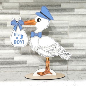 Puede incluir: Decoración de madera de una cigüeña blanca, azul y naranja, que lleva un bebé en un saco azul. El saco tiene un lazo azul y el texto "It's a BOY!". La cigüeña lleva un sombrero azul y una pajarita. Sobre una base redonda de madera.