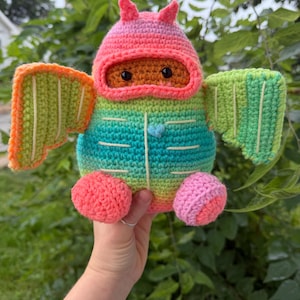 May include: A crocheted plush toy featuring a gradient of green, teal, and orange yarn. The creature has a pink hooded head, two large wings with white stitched lines, a small blue heart on its chest, and round pink and orange feet.
