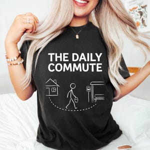 May include: Black t-shirt with the text "THE DAILY COMMUTE" in white. Below the text is a graphic of a house, a stick figure walking with a bucket, and a bus stop.