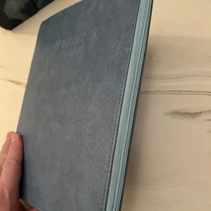 Może przedstawiać: Niebieski notes z zamkiem błyskawicznym z wytłoczonym tekstem "The Gratitude Journal" na okładce.