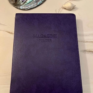 May include: A deep purple magazine folder with the embossed text "MAGAZINE FOLDER" on the front. The folder is made of a textured material. A large, iridescent seashell, a small geode, and a white seashell are in the background.