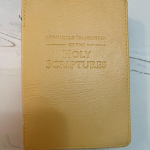 以下が含まれることがあります： 「NEW WORLD TRANSLATION OF THE HOLY SCRIPTURES」の文字がエンボス加工された、タン色の本。ステッチの縁取りがあり、滑らかで革のような質感です。