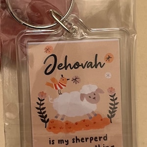 May include: Clear rectangular keychain with a printed design. The design features the word "Jehovah" in script, a cartoon sheep, and the text "is my shepherd I will lack nothing" from Psalm 23:1. A small bird is also included in the design.