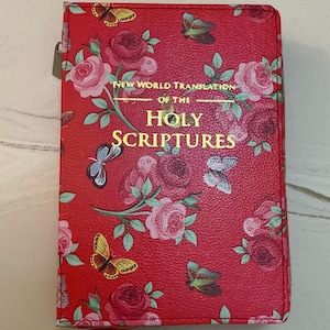May include: A red book cover featuring a floral pattern of pink roses and green leaves interspersed with colourful butterflies. The text New World Translation of the Holy Scriptures is printed in gold lettering on the front centre of the textured cover.